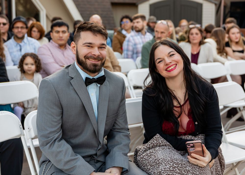 Smiling couple seated at an outdoor wedding ceremony, man in gray suit with bow tie, woman in red top with glittery skirt.