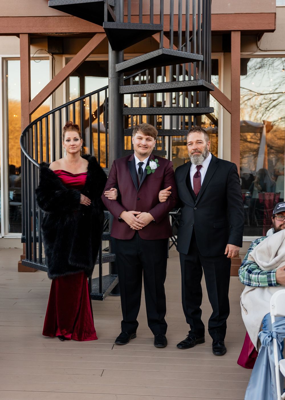 Family of three posing formally outdoors near spiral staircase, wearing elegant attire including burgundy gown and suit.