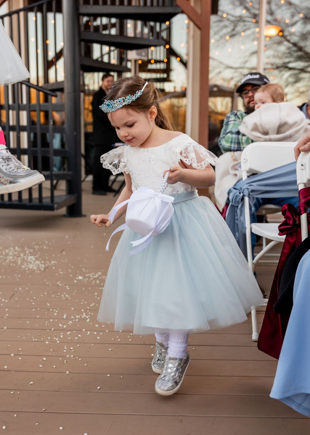 A young flower girl in a light blue tulle dress and tiara scatters petals on a wooden deck at an outdoor wedding.