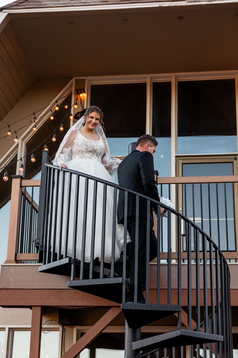 Bride and groom descend outdoor spiral staircase at wedding venue with string lights glowing above.