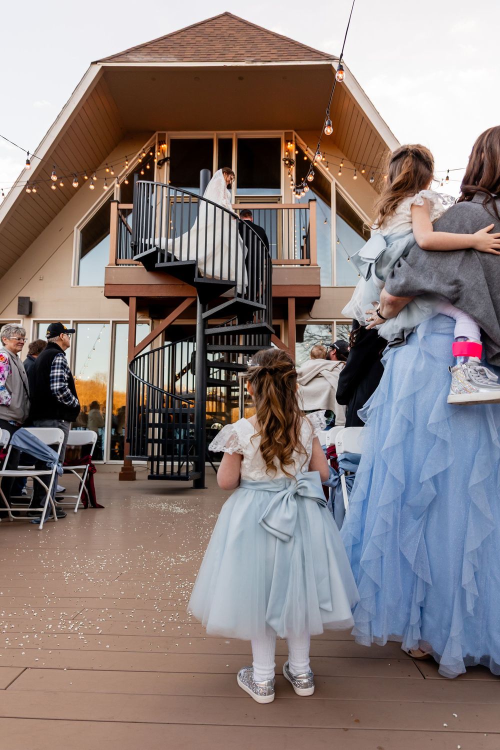 Flower girls in blue tulle dresses watch bride and groom on A-frame cabin deck with spiral staircase at outdoor wedding.
