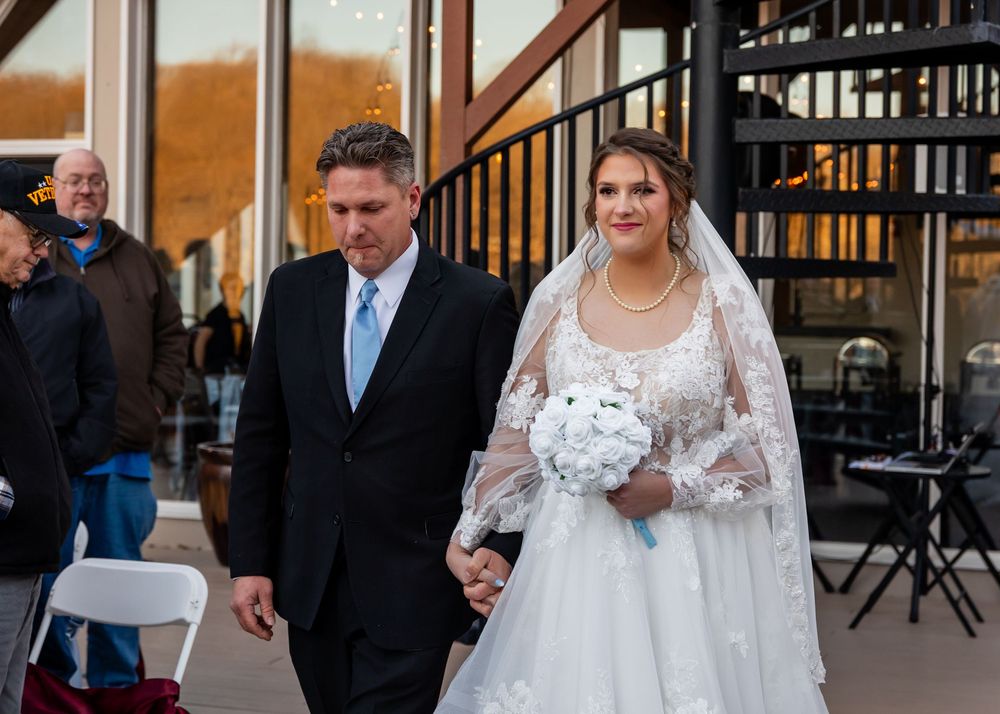 Bride in lace wedding gown holding bouquet walks with suited man outdoors at wedding ceremony venue.