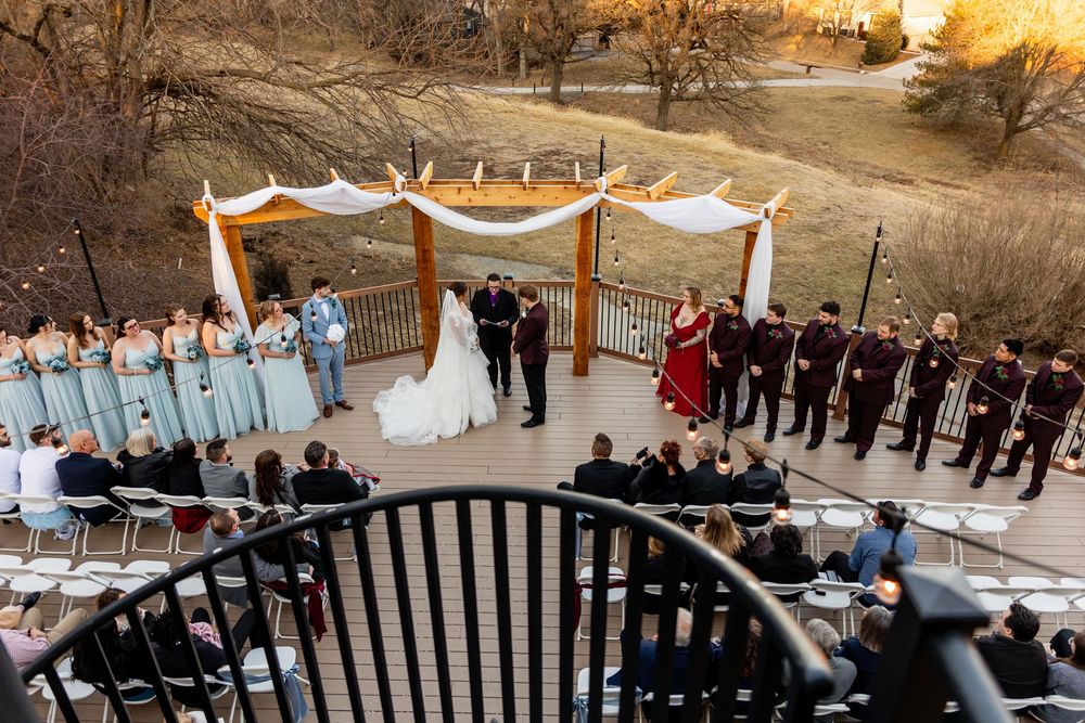 Outdoor wedding ceremony on a deck with bridesmaids in blue, groomsmen in burgundy, and a draped floral arch at sunset.