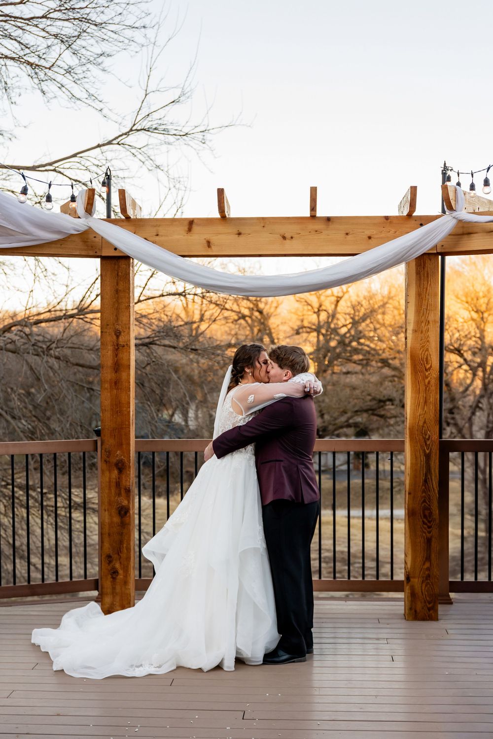 Bride and groom share first kiss under wooden pergola with white draping and string lights on outdoor deck at sunset.