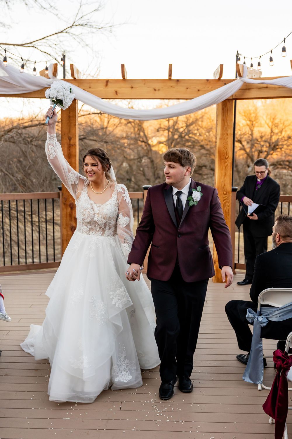 Joyful newlyweds walk down the aisle on an outdoor deck, bride raising bouquet in white gown, groom in burgundy suit.