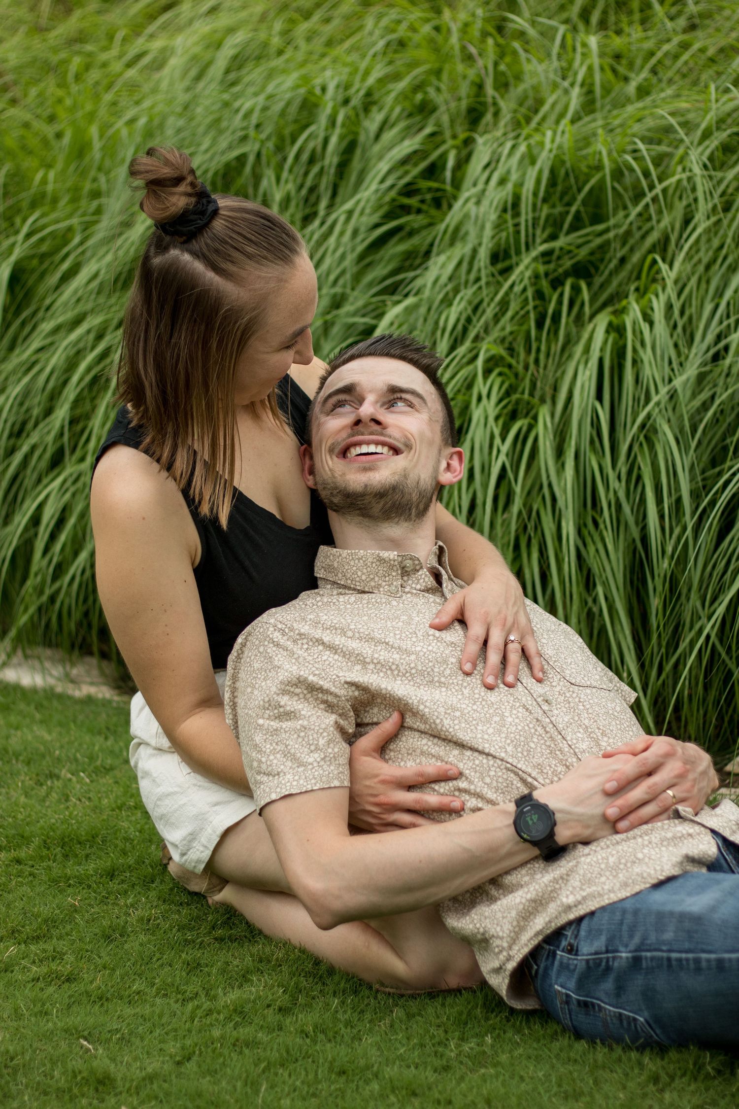 A couple shares an intimate moment together while relaxing on grass with tall reeds in the background.