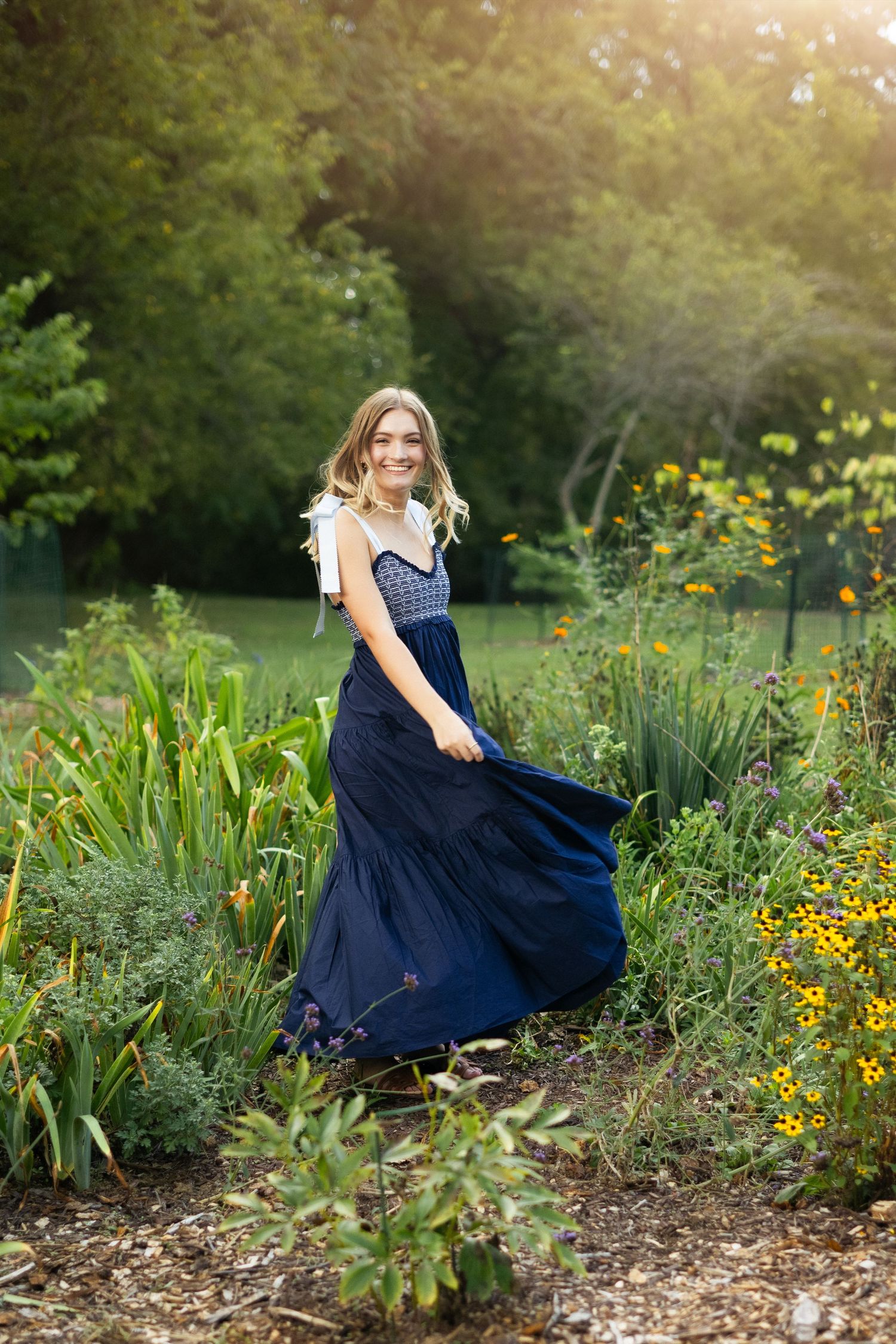 A person in a navy blue dress poses gracefully in a sunlit garden filled with yellow wildflowers and greenery.
