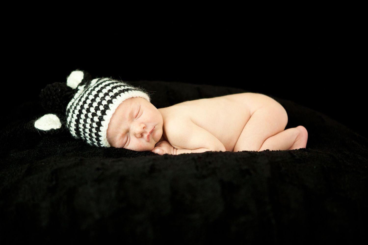 Sleeping newborn baby wearing black and white striped knit hat lying on dark background.