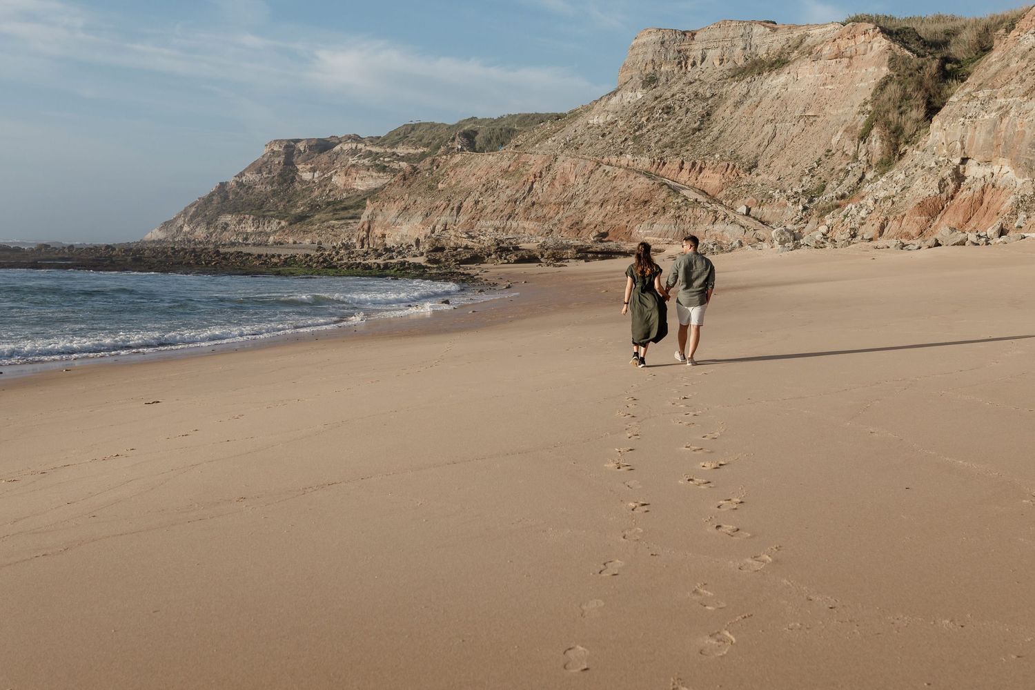 Two people walking along a sandy beach with cliffs in the background.