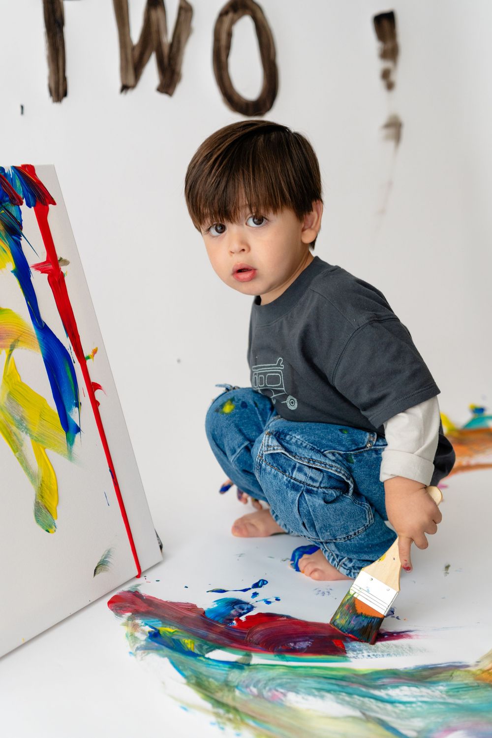 A young boy sitting on the floor and painting with colorful paints on a white background.