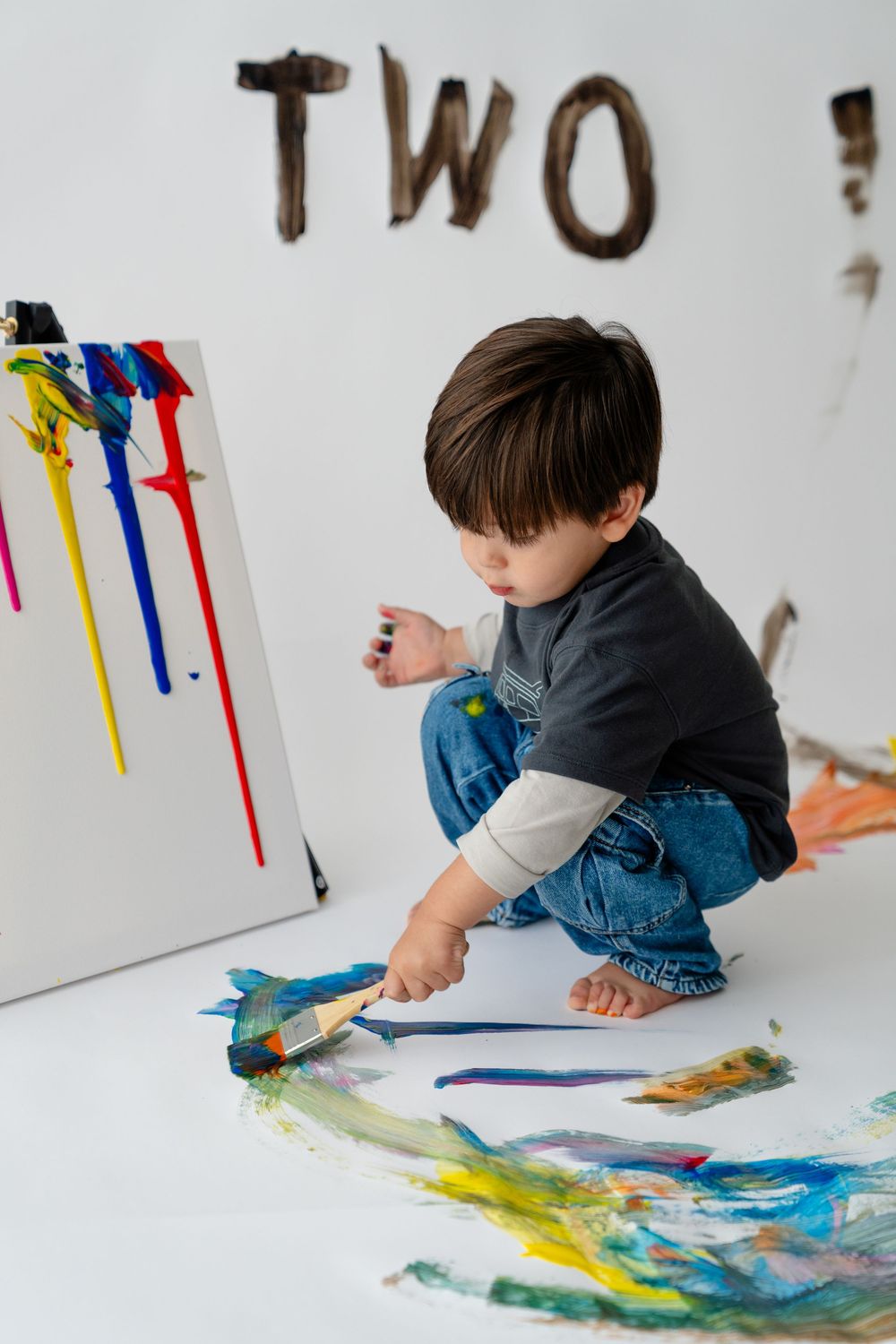 A young boy sitting on the floor and painting with colorful paints on a canvas.