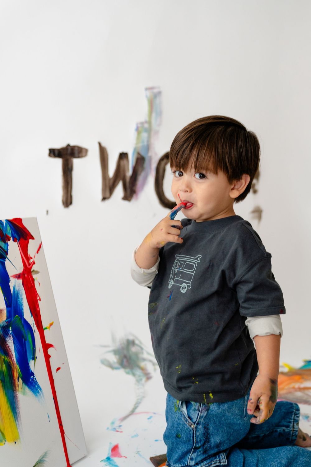 A young child standing in front of a wall with colorful artwork, appearing to be creating their own art.