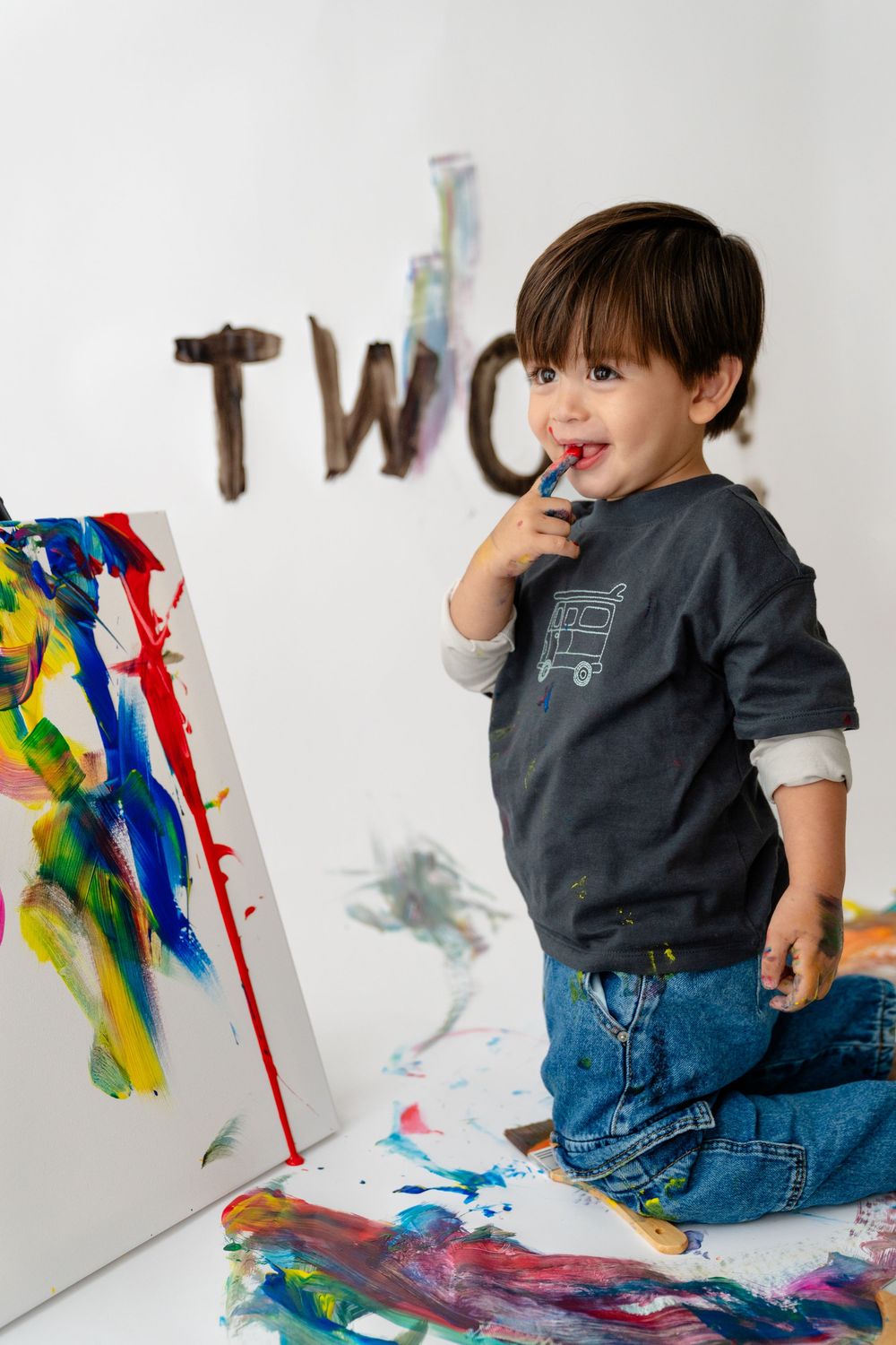 A young boy sitting on the floor, painting with colorful paints and brushes, with the word 'TWO' written on the wall behind him.