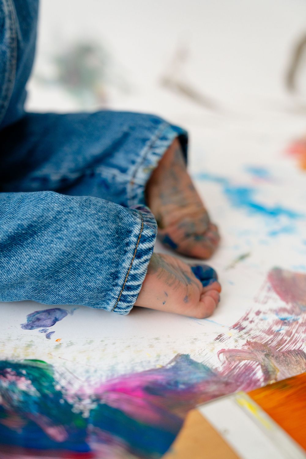 A close-up view of a child's feet and legs, surrounded by colorful paints and art supplies on the floor.