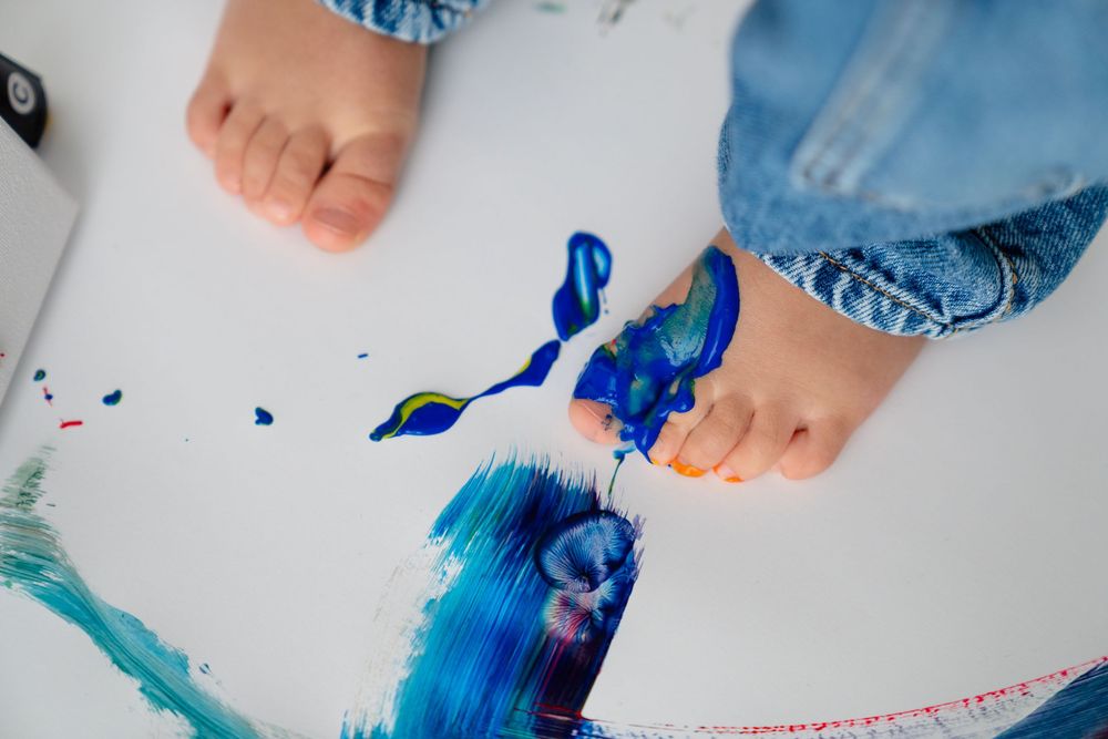 The image shows a person's feet painting with blue and teal colors on a white surface.