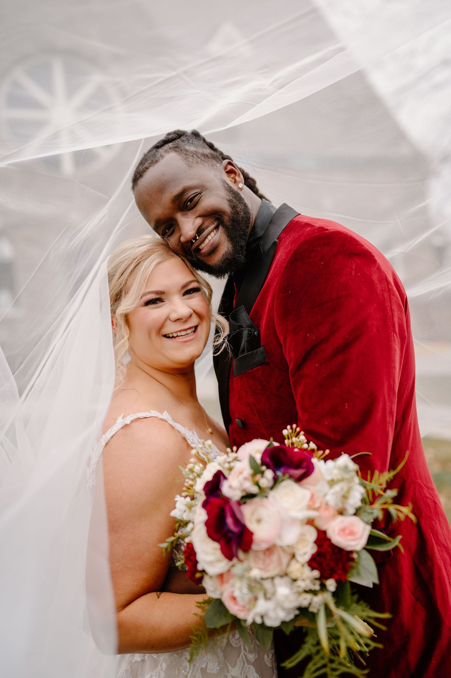 Happy newlyweds share a joyful moment under a veil with a beautiful bouquet of pink and burgundy flowers.