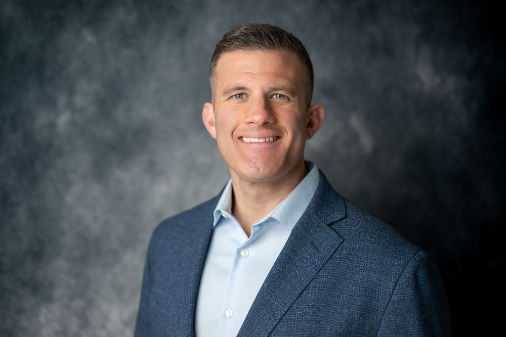 Professional man in a blue blazer and light blue shirt smiling confidently against a textured gray background.
