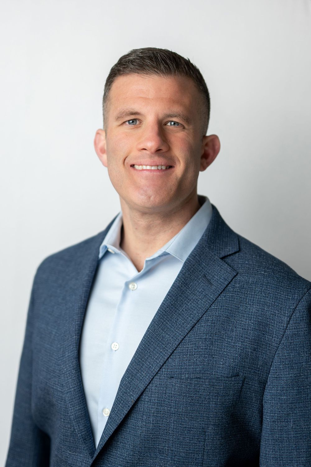Smiling professional man in a blue blazer and light blue shirt posing against a bright white background.