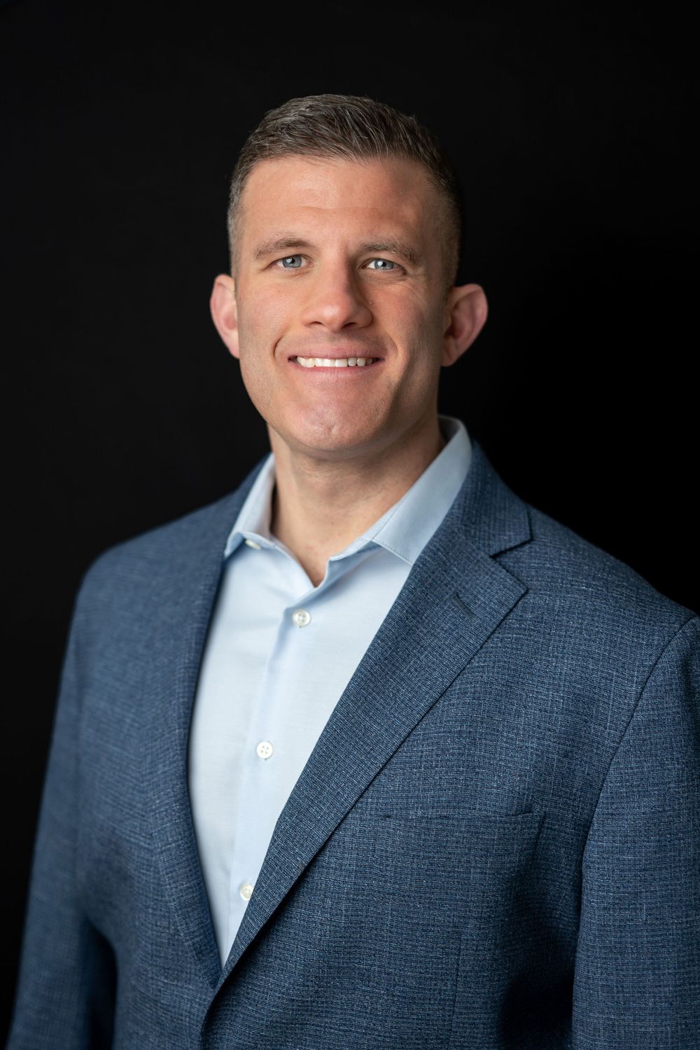 Professional man in a blue blazer and light blue shirt smiling against a dark black background.