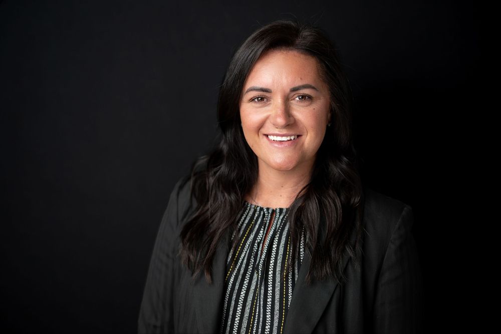 Smiling professional woman with dark wavy hair wearing a black blazer against a dark background.