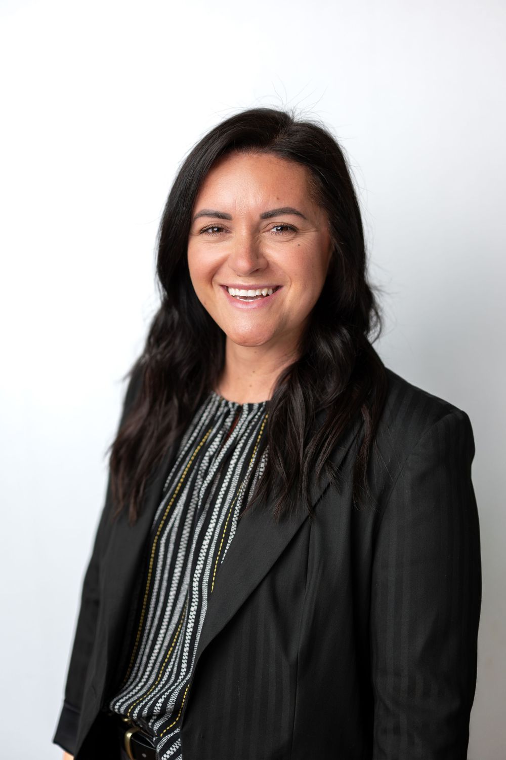 Smiling woman with dark hair in black blazer and striped top, professional headshot on white background.