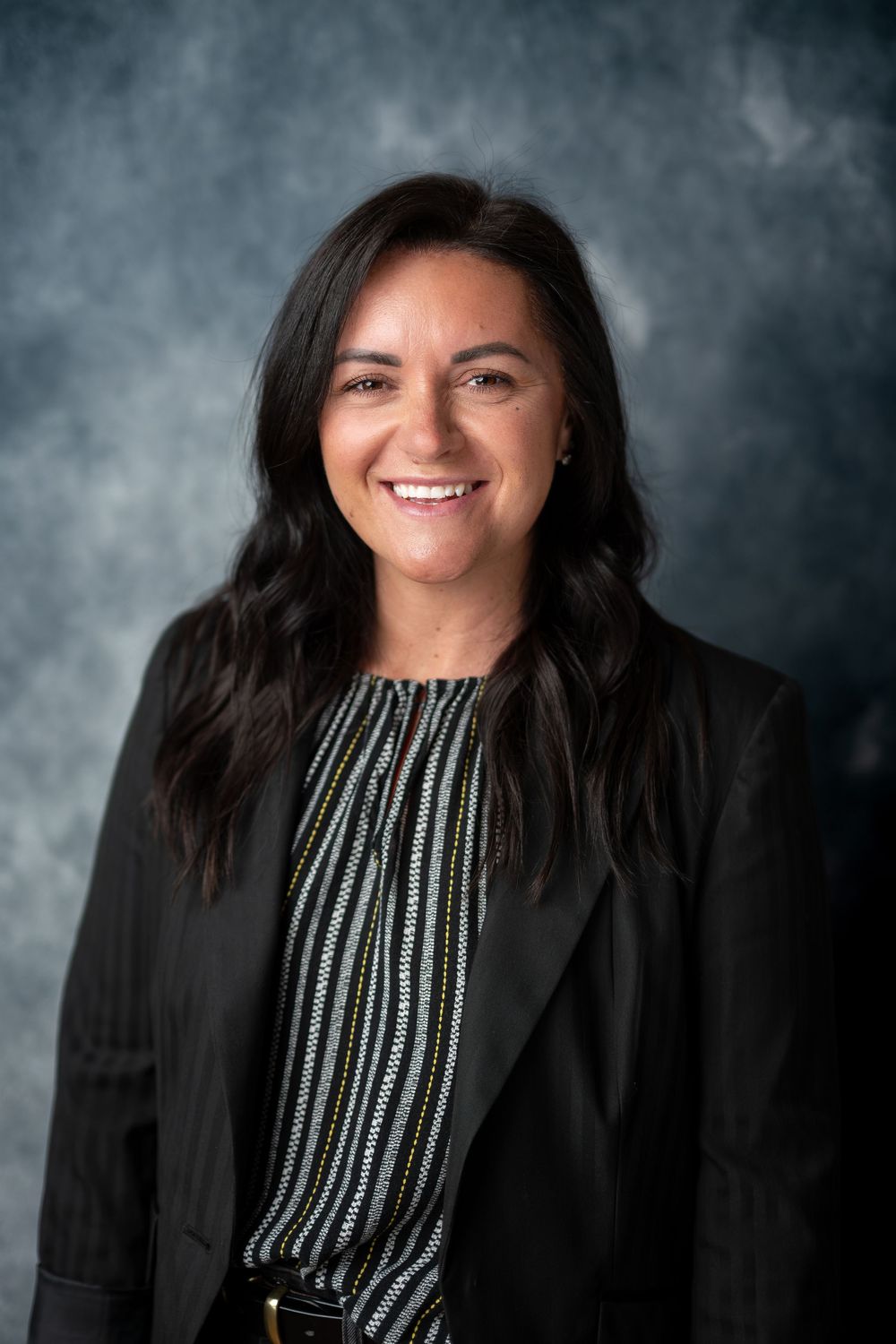 Smiling professional woman with dark hair wearing black blazer and striped top against gray backdrop.