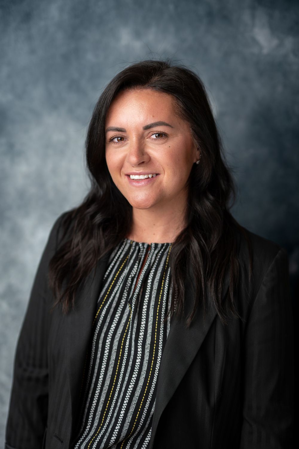 Dark-haired woman in black blazer and striped blouse smiling in professional corporate headshot photo.