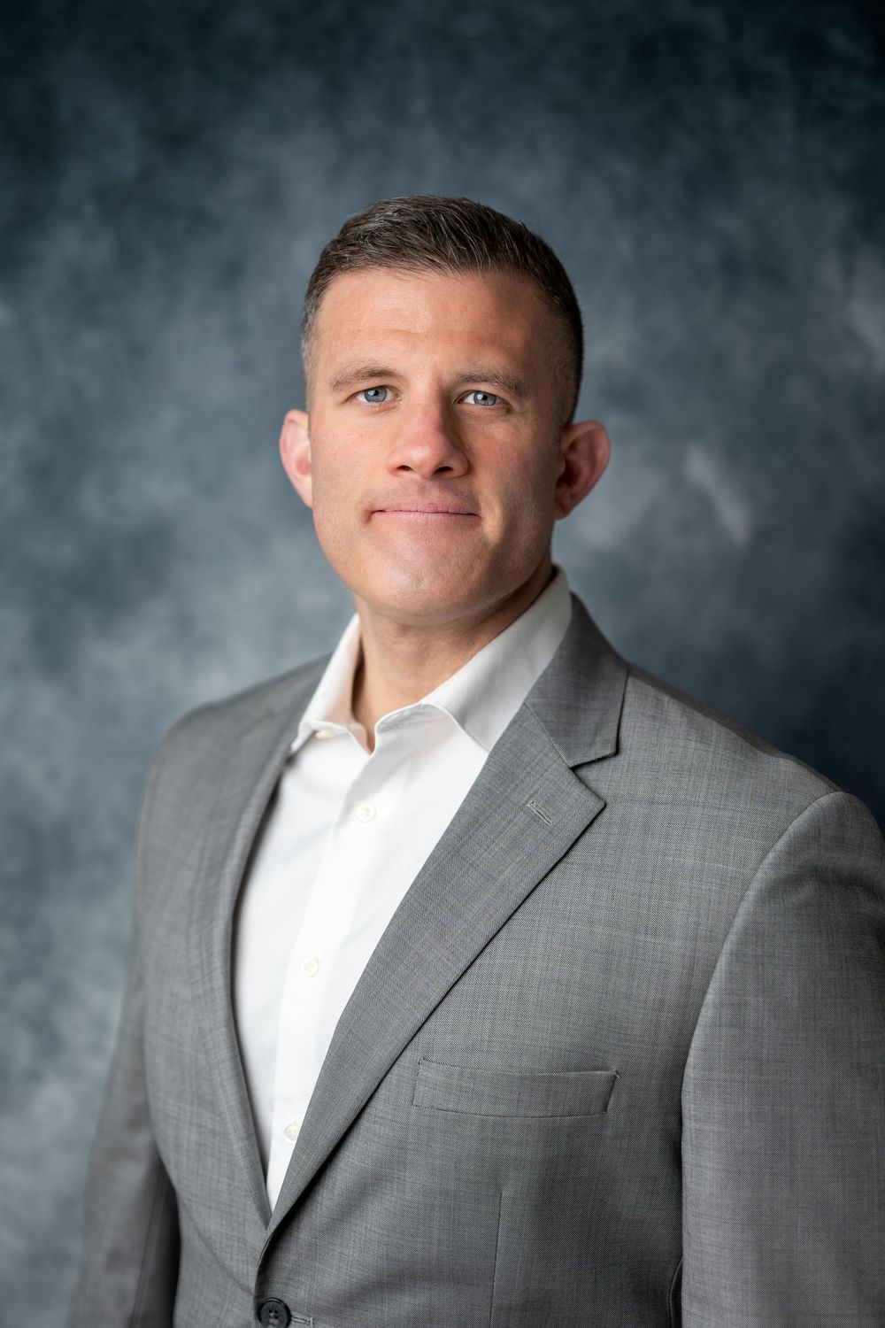 Professional man in gray suit and white shirt with neutral expression against blue-gray studio background.