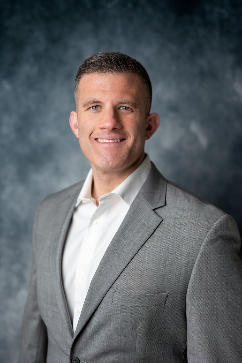 Athletic man with short hair in gray suit smiling confidently in professional studio headshot photo.