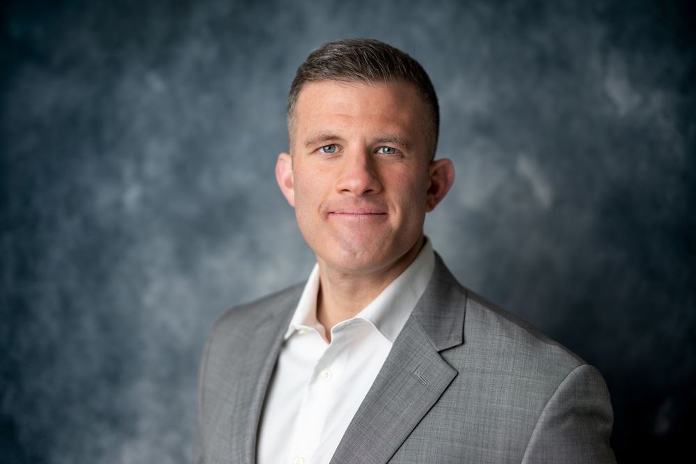 Male professional in gray blazer and white shirt smiling slightly in corporate headshot, dark background.