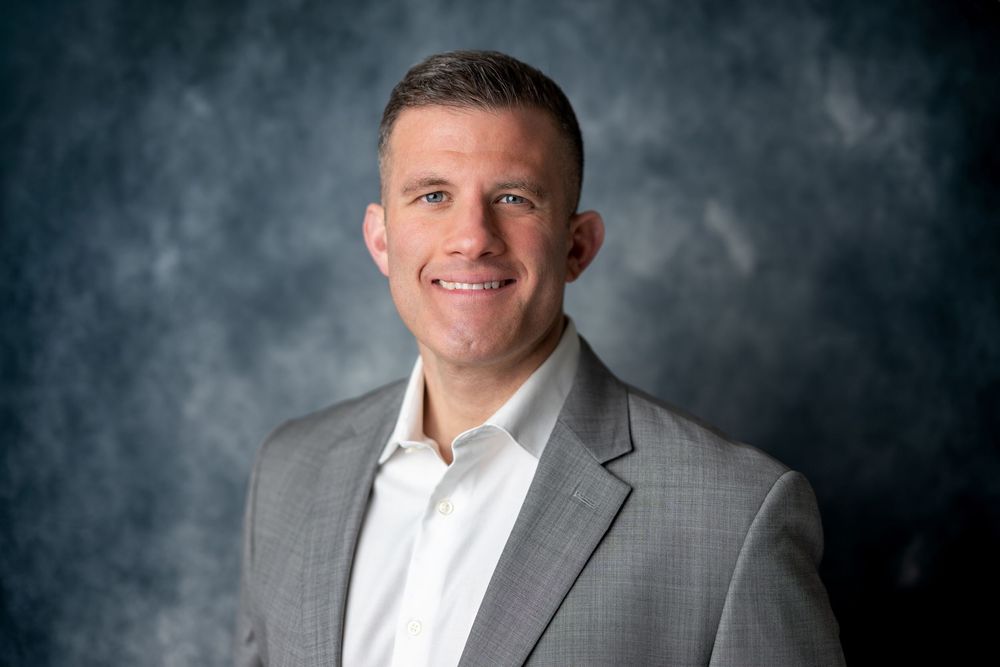 Smiling man in gray suit jacket and white shirt, professional business headshot on gray backdrop.