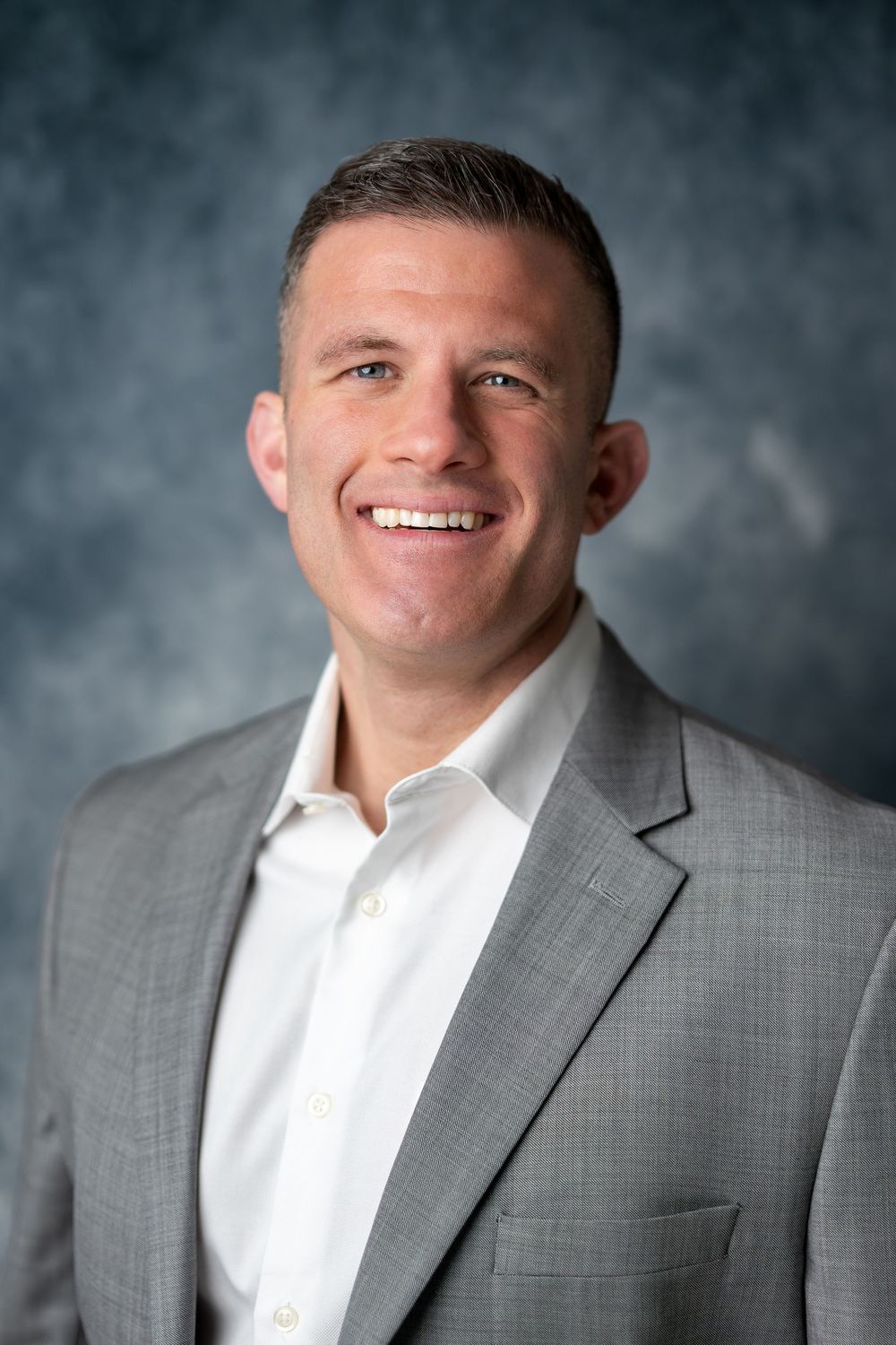 Professional man in gray blazer and white shirt with broad smile against blue-gray studio background.