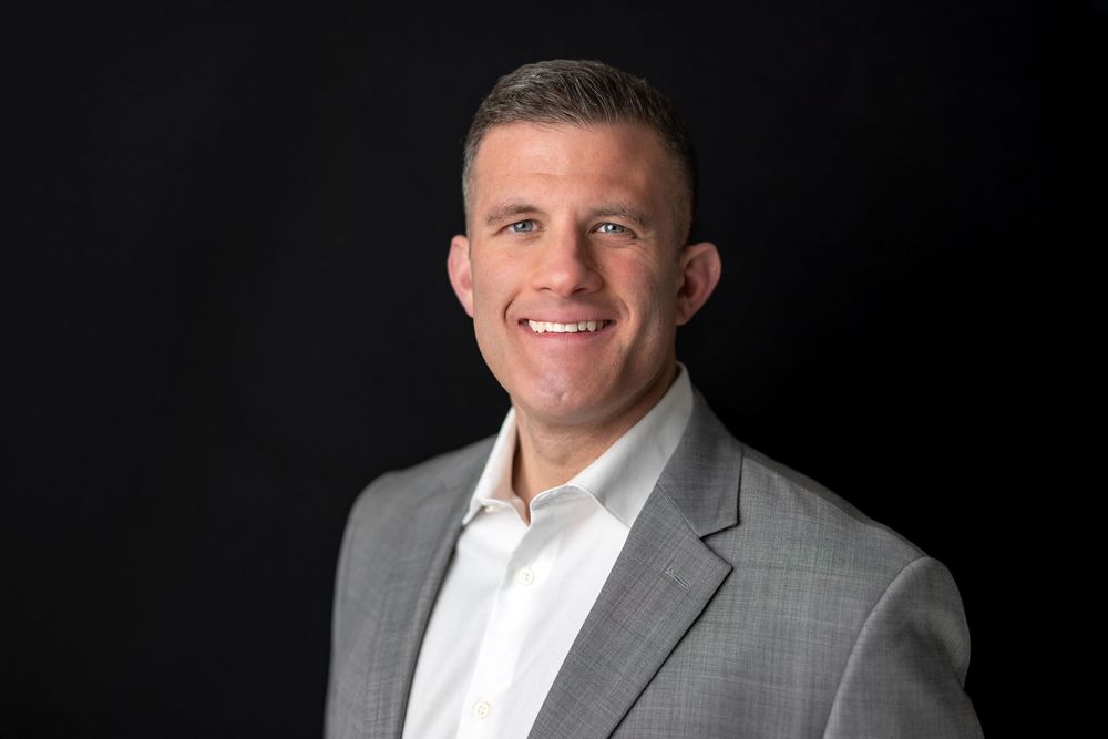 Professional photo of a smiling man in a light gray blazer and white shirt on dark background.