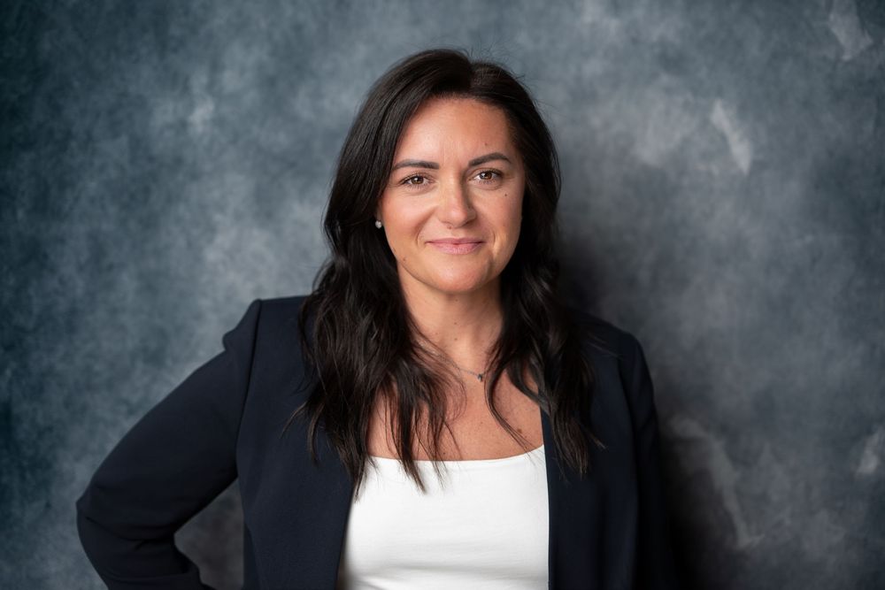 Professional woman with dark wavy hair in a navy blazer and white top posing confidently against a gray background.