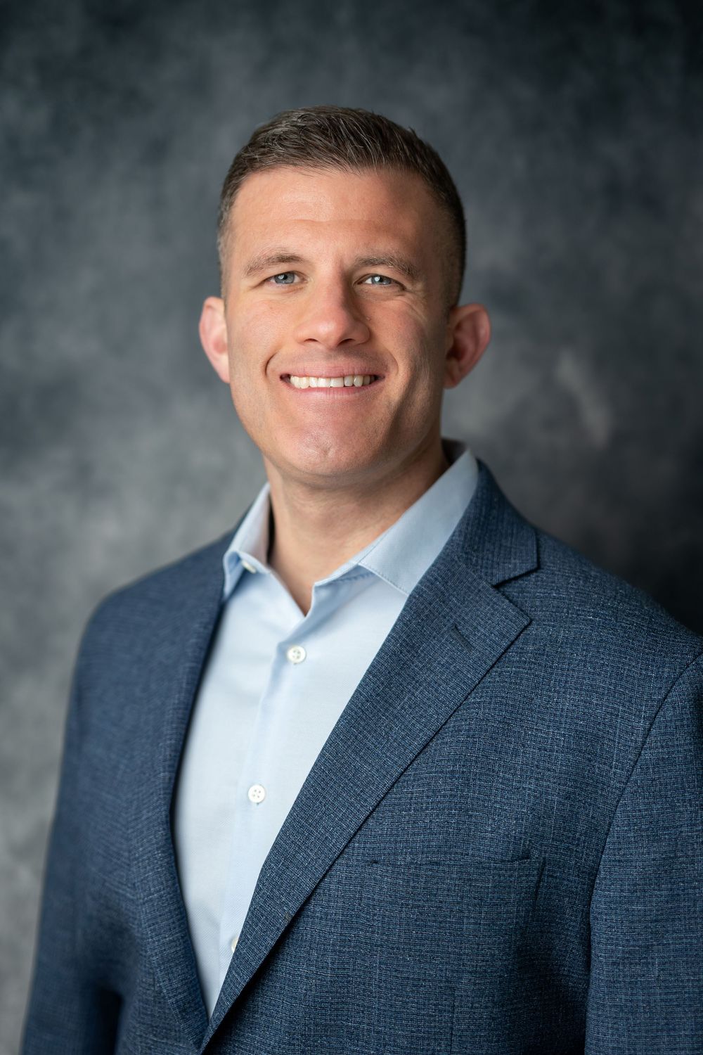 Smiling professional man with short hair wearing a blue blazer and light blue shirt against a gray backdrop.