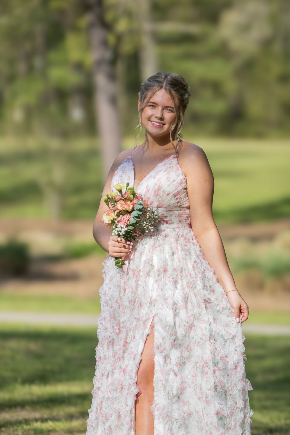 A person in a floral white dress poses outdoors in a sunny garden setting holding a bouquet of flowers.