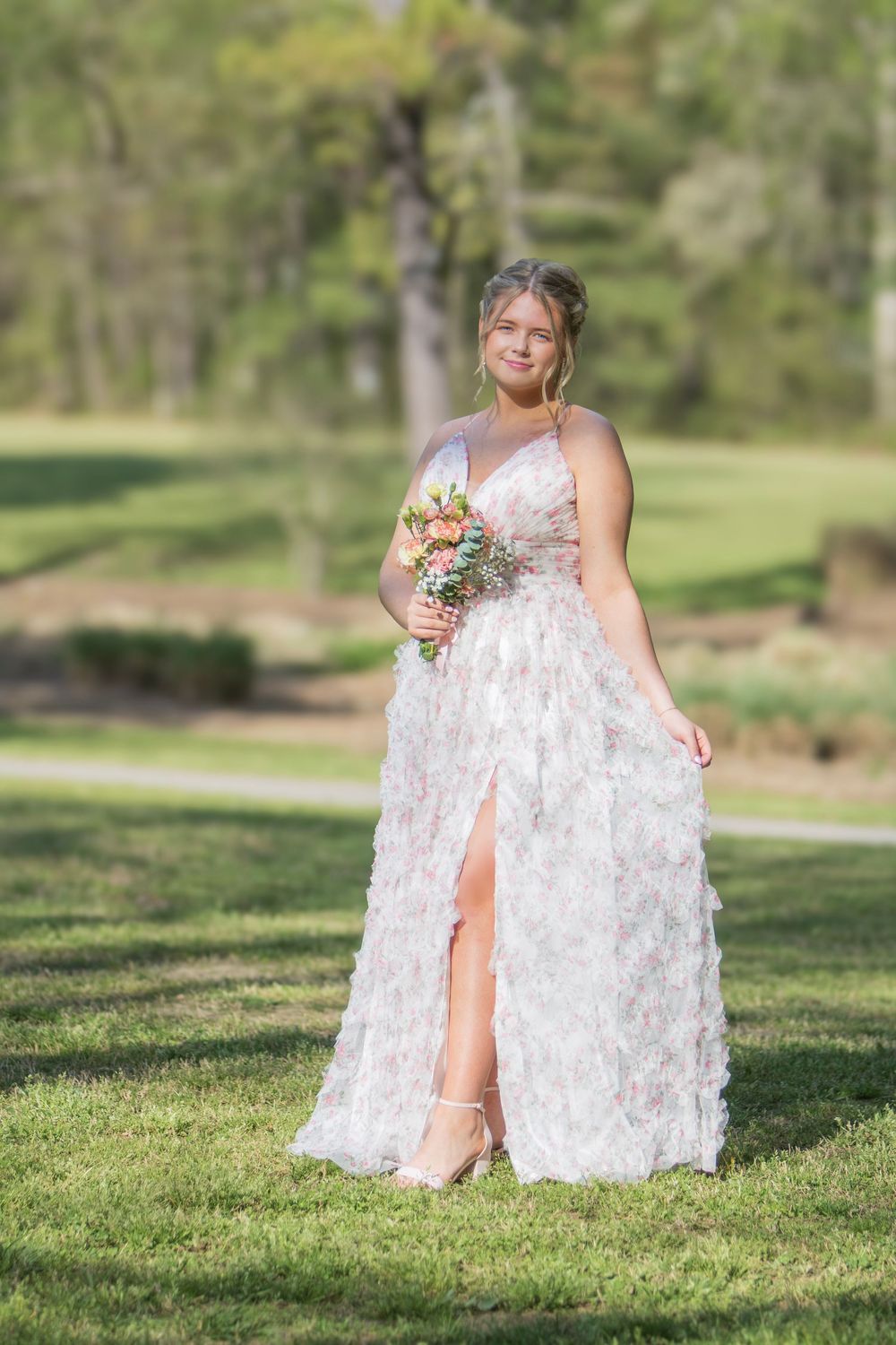 A person in a white floral wedding dress poses gracefully on grass in a sunny outdoor garden setting.