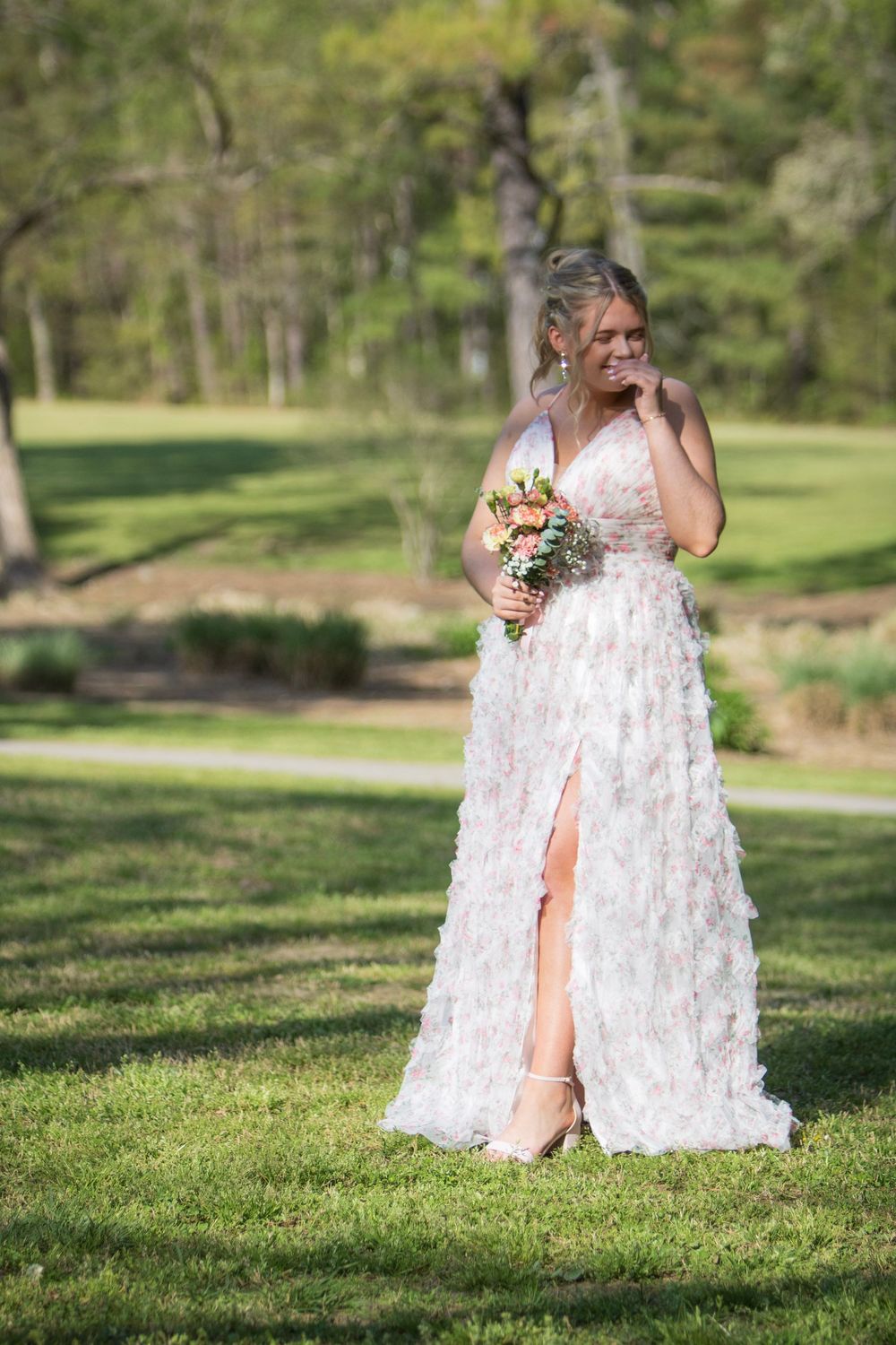 A white wedding dress with floral appliques flows elegantly across a sunlit green lawn in a garden setting.