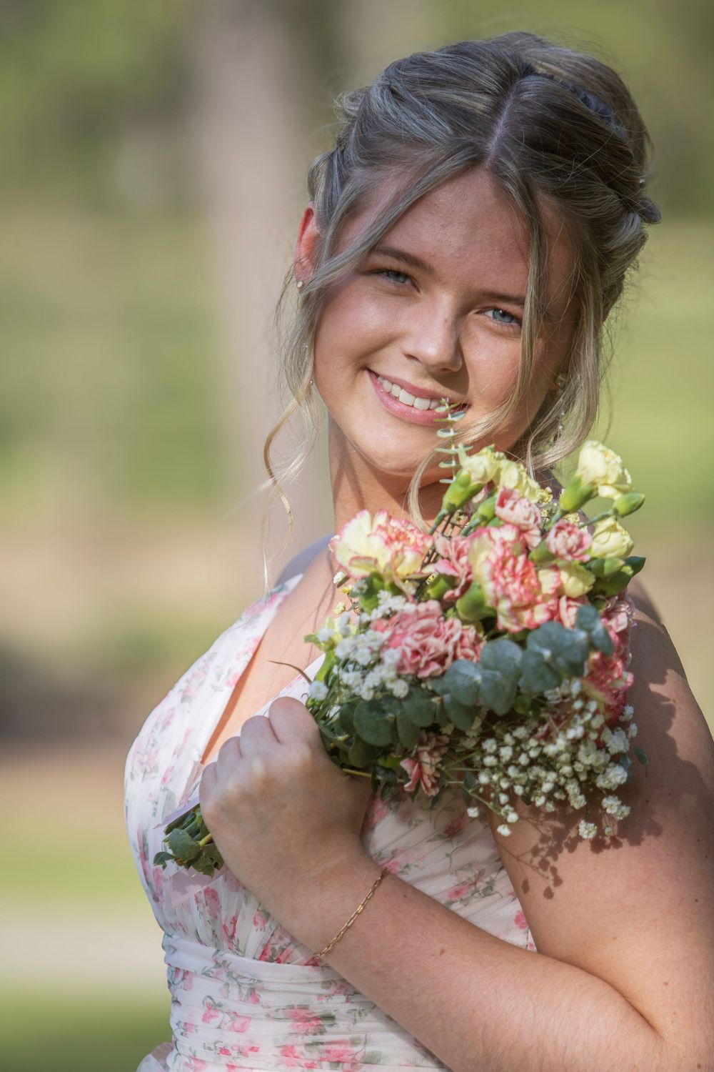 A person in a floral dress poses outdoors with a bouquet of pink and white flowers in a natural setting.