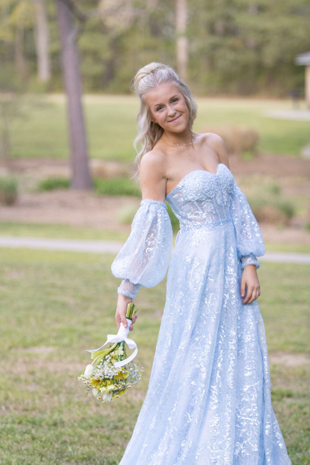 A person in a flowing light blue lace dress poses gracefully in a sunny outdoor garden setting.