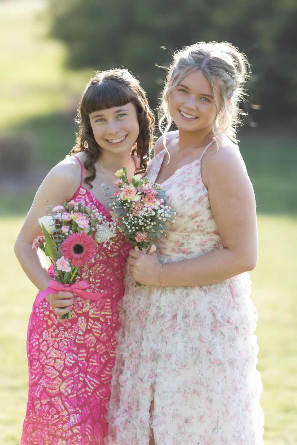 Two people wearing pink and white floral dresses embrace and smile while holding bouquets in a sunny outdoor setting.