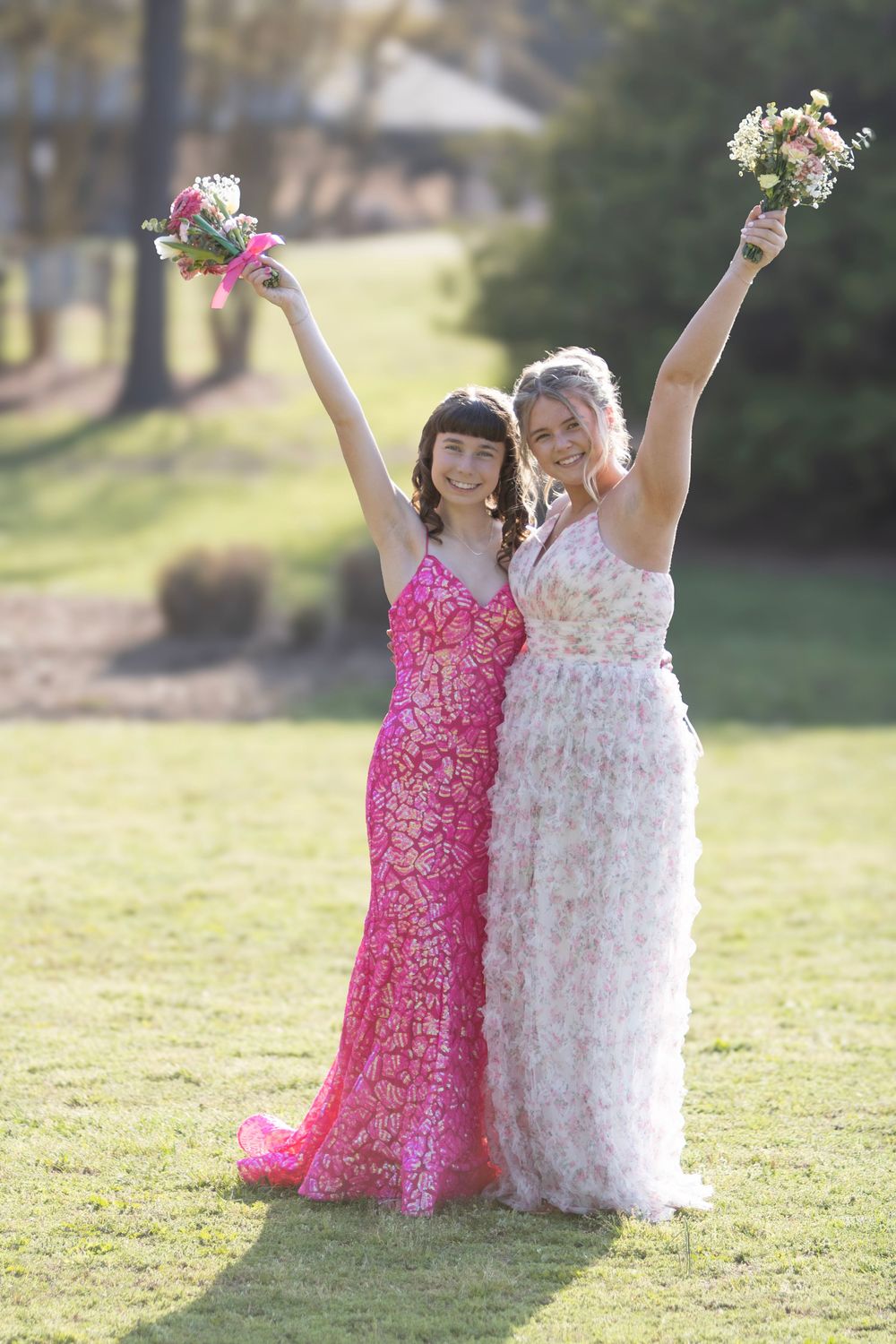 Two young girls in flowing pink and white dresses dance together joyfully on a sunny lawn with raised bouquets.