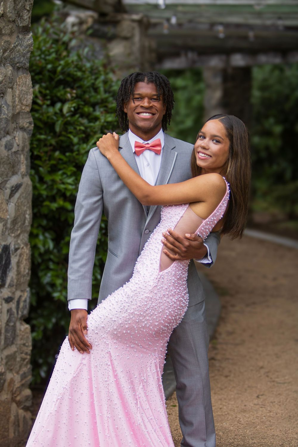A couple in formal attire, with a pink dress and grey suit, poses playfully in a wooded outdoor setting.