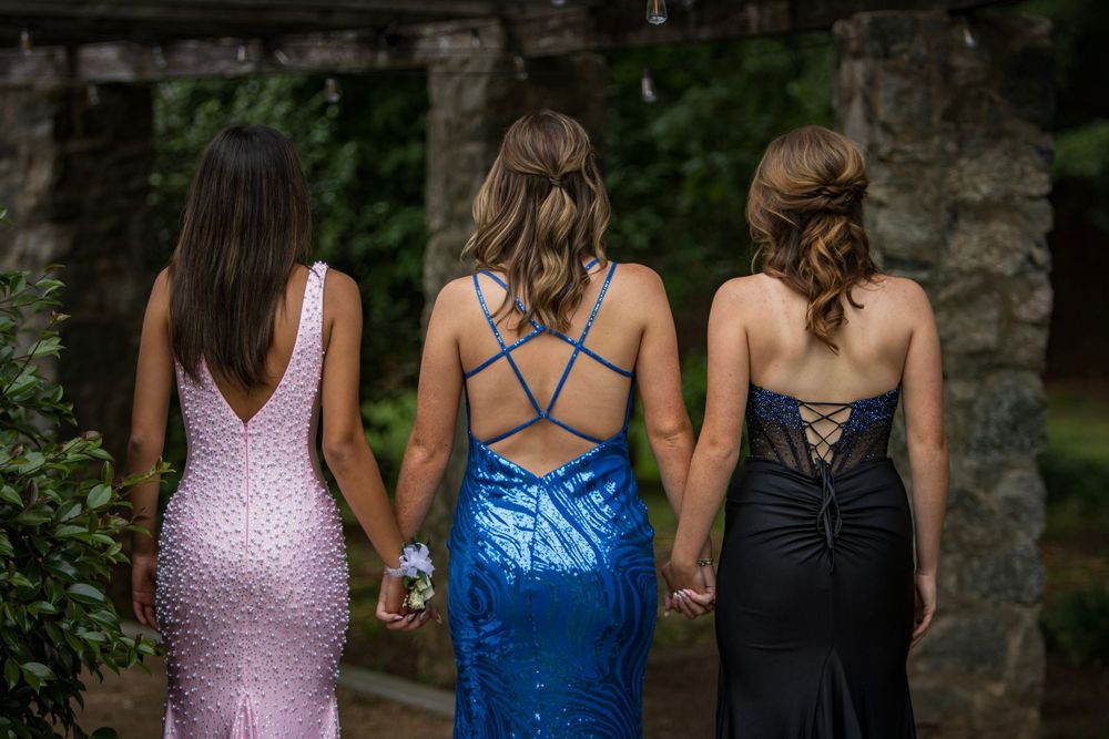 Three women in formal evening gowns - pink, blue and black - holding hands while facing away from the camera.