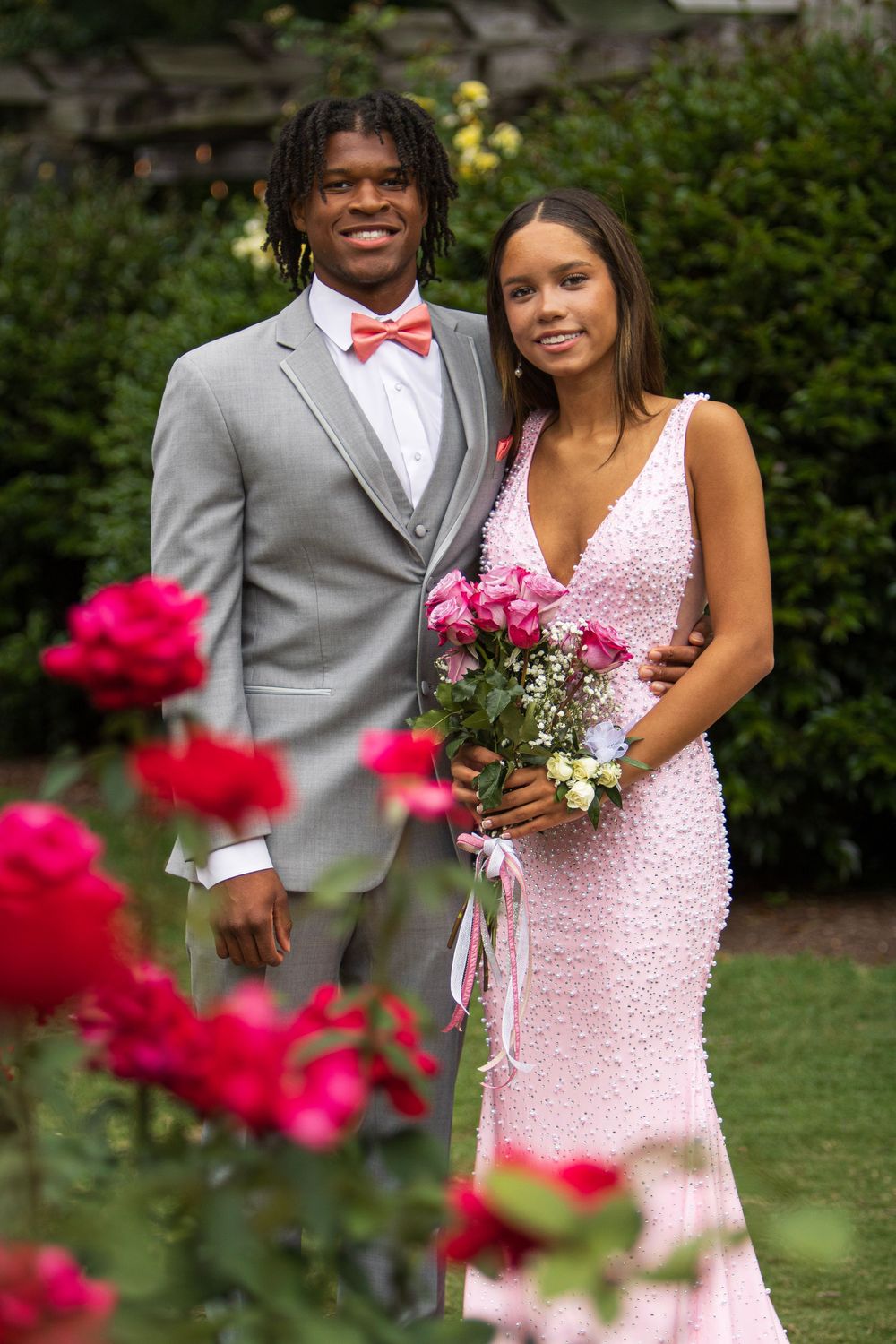 A couple in formal attire poses together at an outdoor wedding, with the bride in a pink dress and groom in grey suit.