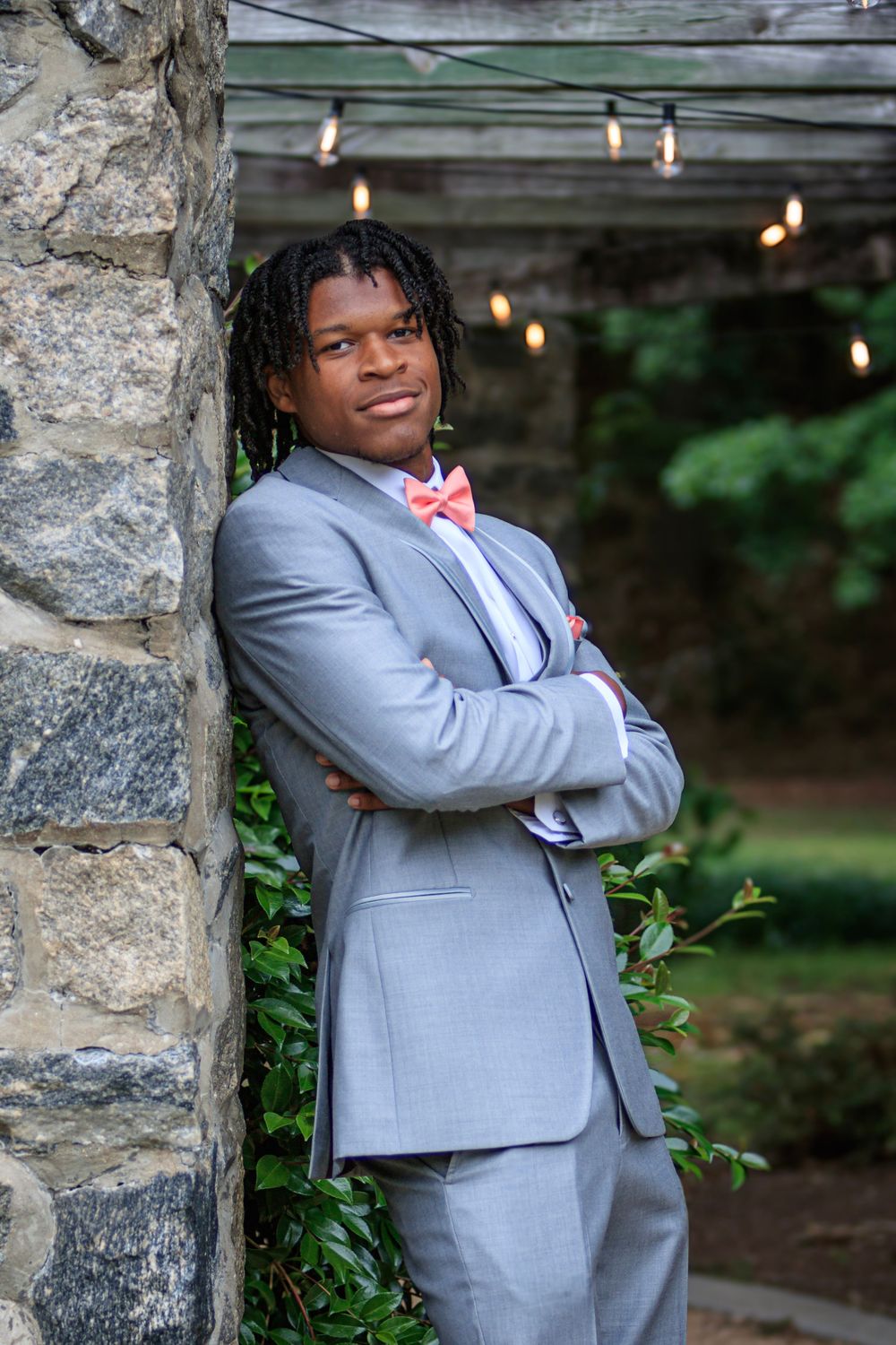 A person wearing a light gray suit and red tie poses confidently against a stone wall with string lights in the background.