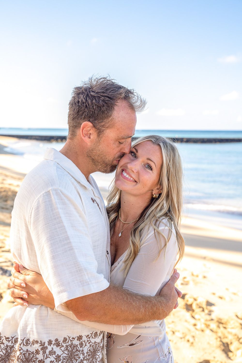 A couple embraces lovingly on a sunny beach with ocean waves and sand in the background.