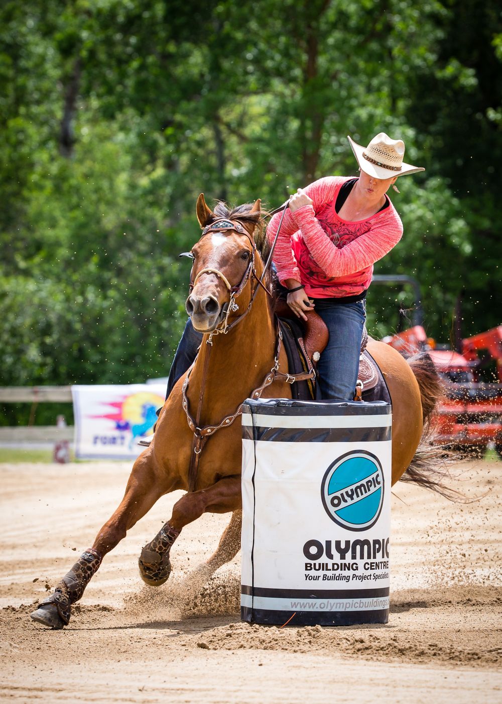 Barrel racing competitor on brown horse rounding a white Olympic building supplies barrel at equestrian competition.
