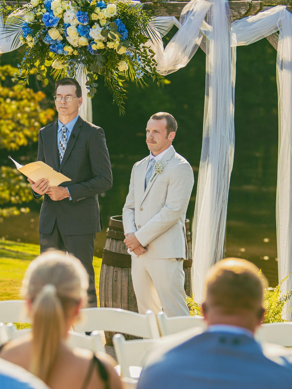 Outdoor wedding ceremony under a white draped arch with floral decorations and guests seated in rows on a sunny day.