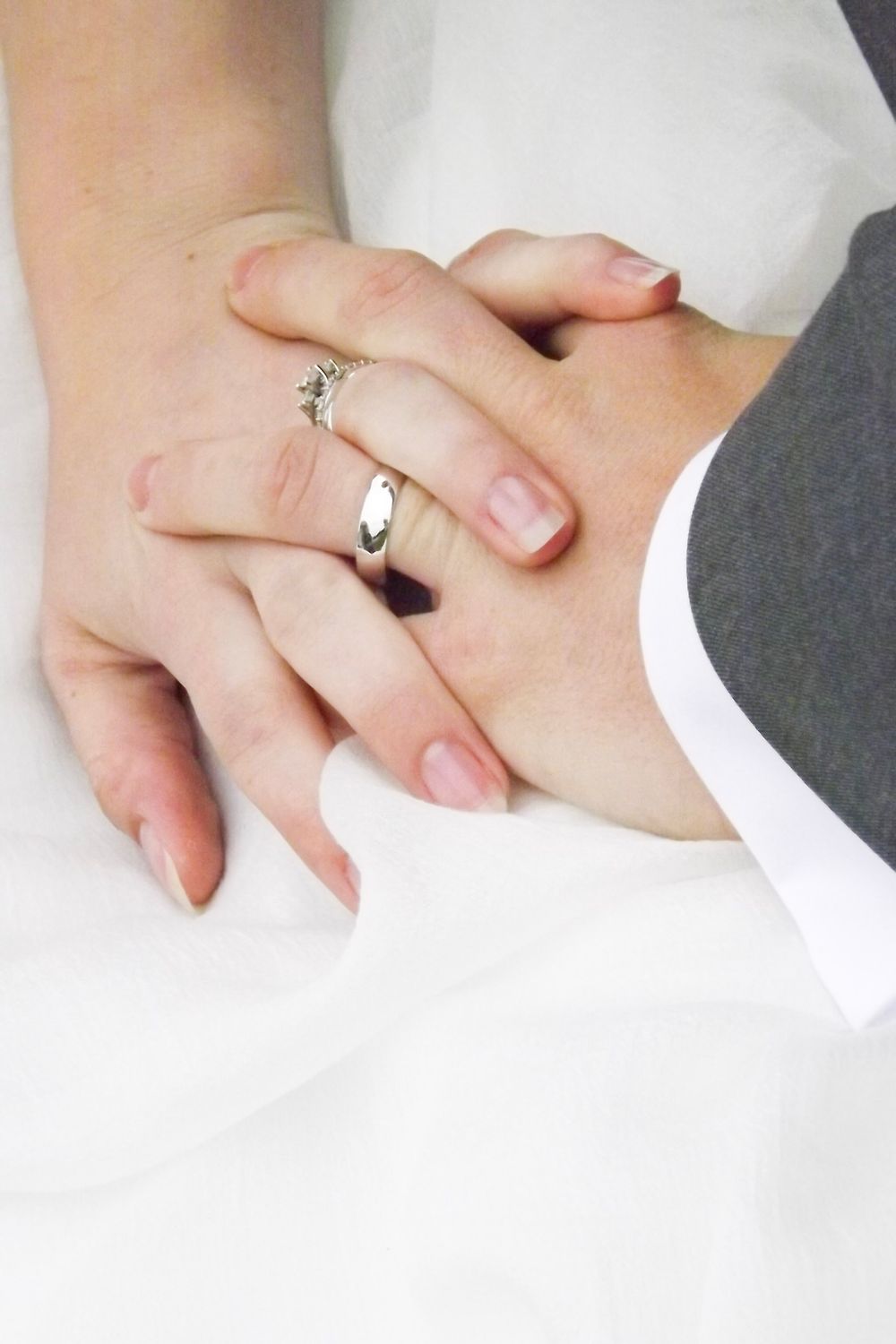 Close-up of two hands with wedding rings touching tenderly against white and gray formal attire.